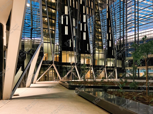 Image of a modern building with contemporary architectural design, featuring glass walls and a steel structure with slanted frames. The building is seen in the evening with soft lighting highlighting its details. In the foreground, there is an open area with some trees and plants arranged in an orderly manner.