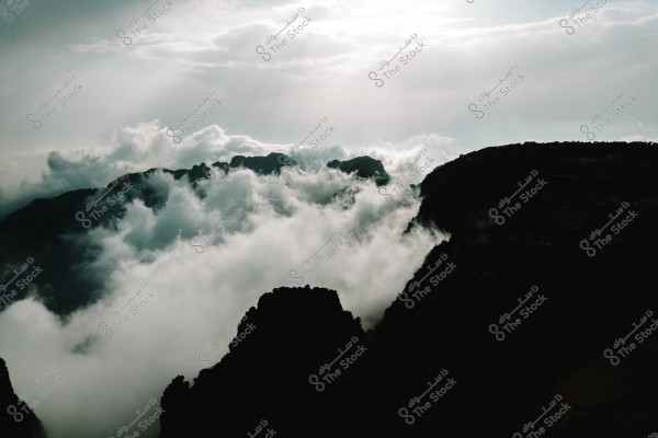 A natural landscape of mountains covered with dense clouds, with sunlight peeking through the clouds in the sky. The scene appears calm and majestic, with a clear contrast between the dark mountain peaks and the white clouds.