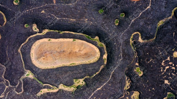 صورة جوية لمنطقة طبيعية تظهر تضاريس فريدة من الصخور البركانية الداكنة المحاطة بالرمال. يوجد في منتصف الصورة تشكيل صخري بيضاوي الشكل مع حواف واضحة, وتنتشر بعض المساحات الخضراء بشكل غير منتظم في الصورة.