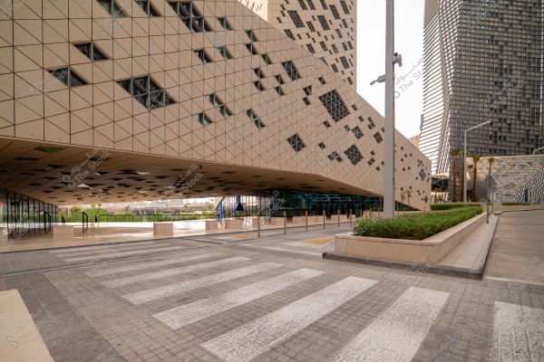 Image of a modern architectural structure featuring buildings with artistic geometric design, characterized by triangular panels and slanted walls with scattered small windows. In the foreground, there\'s a street with green seating areas and a pedestrian walkway. In the background, tall buildings with a similar design reflect a distinctive urban character.