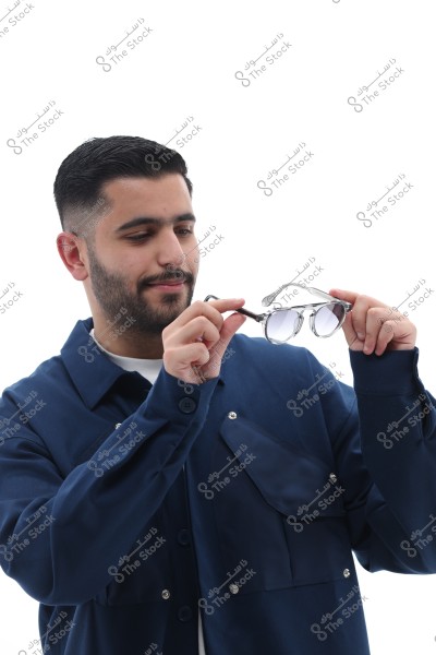 The image shows a person wearing a blue jacket holding a pair of sunglasses in their hands. The background is white, and the person appears focused on the sunglasses.