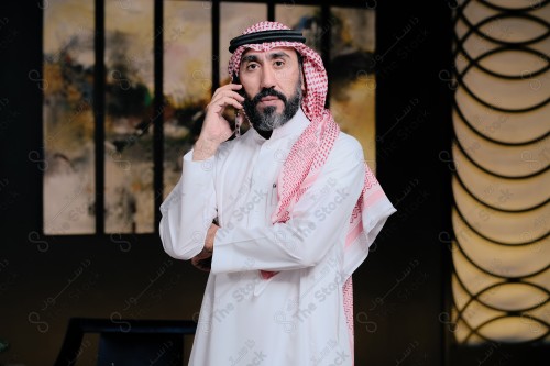 رجل سعودي يرتدي الثوب و الشماغ، يتفاعل وينظر الى الكاميرا وهو يتحدث عن طريق الجوال