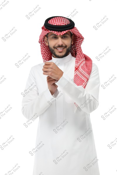 A portrait of a man wearing traditional Saudi attire, consisting of a white thobe and a red and white ghutra with a black agal. The man is smiling and adjusting the sleeves of the thobe, with a completely white background.