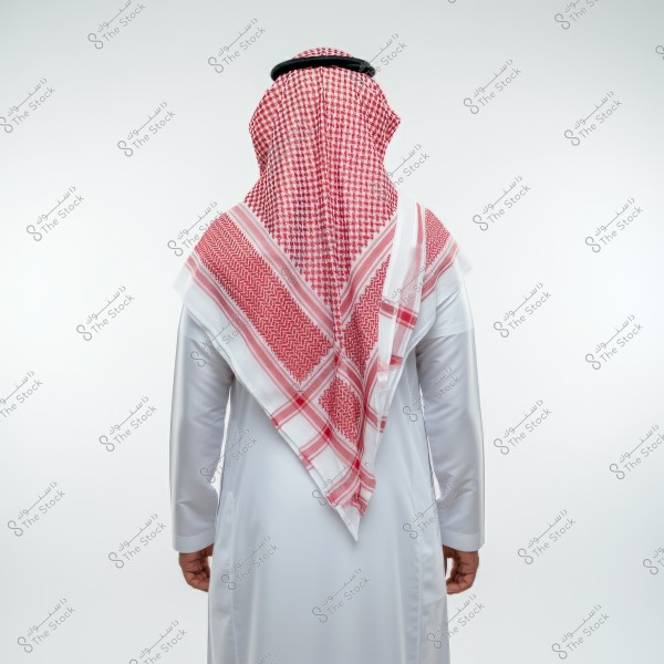 The image shows a man standing from the back wearing the traditional Saudi white thobe and a red and white checkered headscarf, known as a ghutra, with a black cord called agal. The background is white.