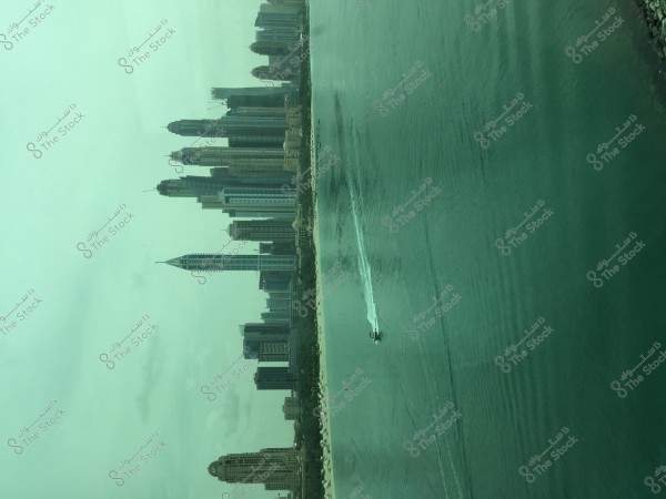 A view of a modern city featuring a collection of tall skyscrapers overlooking the sea. The buildings have sleek and contemporary architectural designs, and the city skyline is clearly visible. In the foreground, a speedboat is moving through the green waters, leaving a wake behind as it heads away from the city.