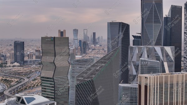 An image of the main financial district in Riyadh, Saudi Arabia, showcasing a collection of modern and diverse architectural buildings. The scene includes tall and distinctive towers, including a uniquely designed tower on the left resembling a staircase, with the cityscape extending under a cloudy sky.