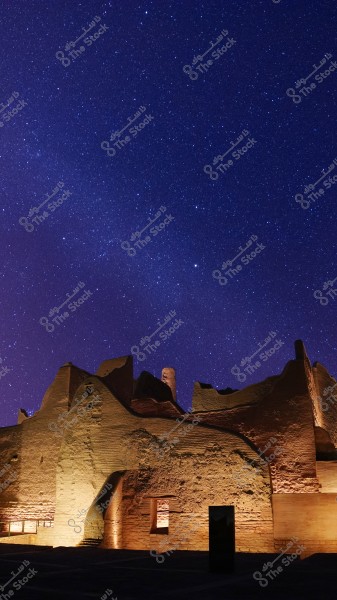 An image of an illuminated archaeological site at night under a starry sky. The ancient mud-brick structure displays its architectural details majestically with contrasting lighting, and the dark blue sky is filled with sparkling stars.