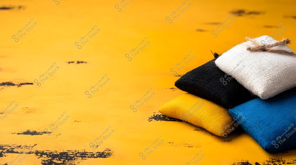 A set of small colored cushions stacked on top of each other on a yellow surface. The cushions are blue, black, yellow, and white, covered with textured fabric, and the white cushion is tied with a coarse rope.