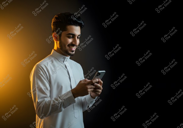 A portrait of a man smiling while looking at his mobile phone. He is wearing a traditional white thobe and standing against a black background, with warm yellow lighting casting a glow on his left side.