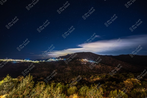 A nighttime image of a natural scene featuring a star-filled sky over a mountainous area. The picture shows hills covered with dense vegetation and village lights illuminating the lands in the distance. Light clouds drift across the sky, adding beauty to the nighttime view.
