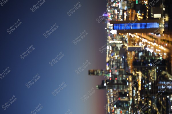 A nighttime image of a city illuminated by electric lights, showing tall buildings from a side view. The sky is dark, indicating night, and there are many bright, colorful lights on the buildings and streets.
