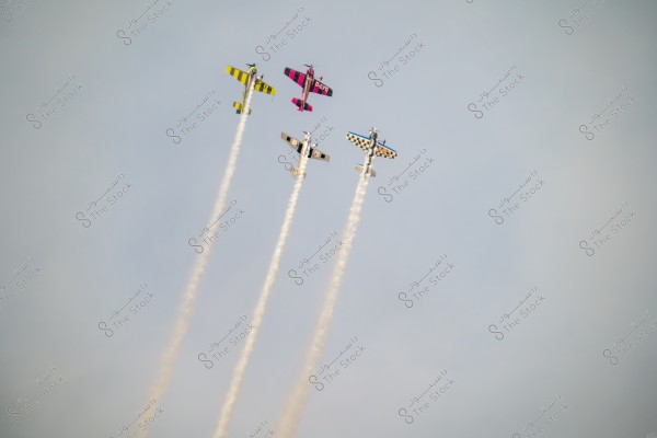 Four stunt planes flying in the sky, tilted slightly to the left, leaving smoke trails in the air. The planes are brightly colored, featuring yellow, pink, and blue with various patterns.