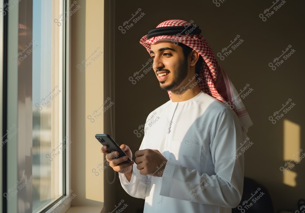 Portrait of a man wearing a traditional Gulf white thobe and a red-checkered ghutra secured with a black agal. He is standing near a window in a sunlit room, holding a mobile phone and smiling, with the sunlight illuminating his face.