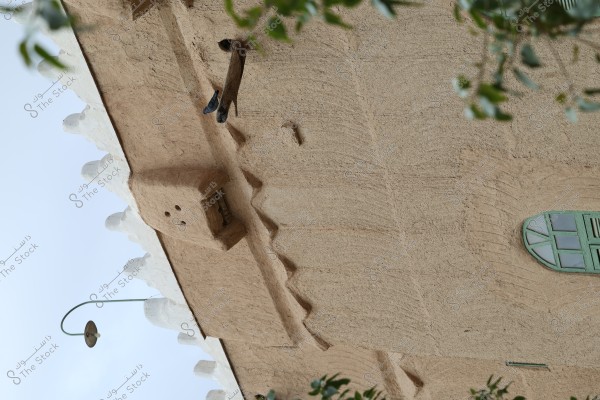 Image of a traditional mud-brick building. The roof features white-edged and protruding decorations. There is a rectangular green window on the wall, along with a hanging lamp from the roof. Some green branches appear at the top.