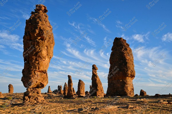 Large natural rock formations in a desert region under a clear blue sky with wispy clouds. The rocks appear to be precariously balanced on their bases, creating a landscape reminiscent of natural sculptures. The ground is dry and sandy.