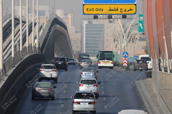 The image shows busy traffic on an overpass in a city with tall buildings in the background. A blue and yellow road sign reads \"Exit Only\" in both Arabic and English. Various types of cars and sizes are moving in two lanes, with a bus visible ahead. The architectural design of the roads and bridges reflects a modern urban style.