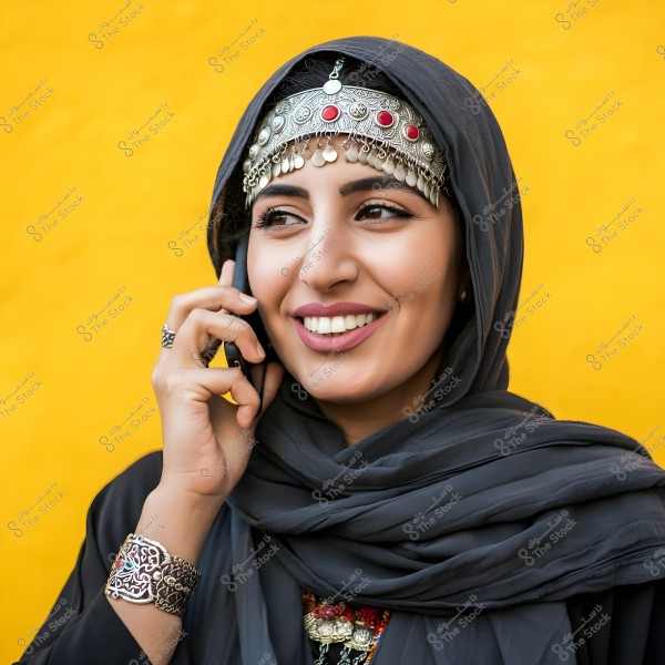 A portrait of a woman wearing a gray scarf and an ornate silver headpiece with red stones, speaking on a phone. The background is a vibrant yellow, and she is smiling while wearing a decorative silver bracelet.