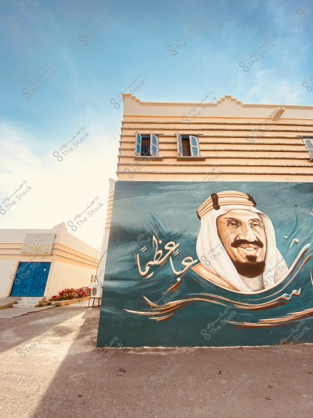 The image shows the side wall of a building with a mural of a man wearing traditional Arab attire, including a thobe, agal, and ghutra. Above the mural are two open windows, and the sky is clear in the background. The shadow of the building stretches onto the pathway leading to a decorative blue door. Red flowers are planted next to the door.