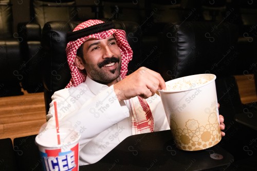 شاب سعودي يرتدي الزي السعودي التقليدي يستمتع بمشاهدة الفيلم أثناء تناوله الفشار في صالة السينما