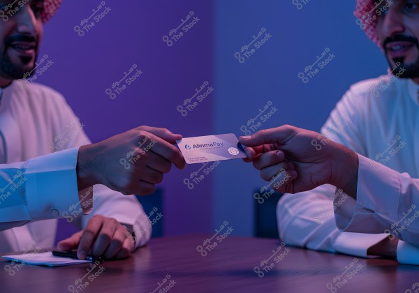 An image of individuals wearing traditional Saudi attire, including a thobe and ghutra, sitting around a table in soft blue lighting. A business card with \"alimmapay\" is being exchanged, indicating a business interaction or meeting.