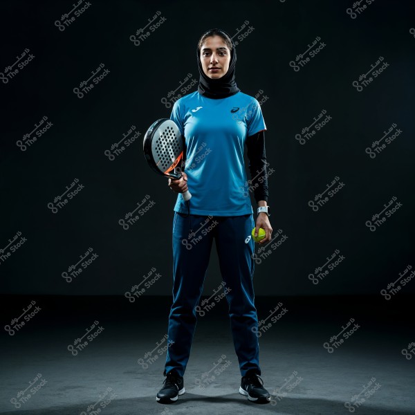 A sports portrait of a woman wearing a hijab and blue athletic attire. She is holding a padel racket and a small yellow ball. She stands on a dark gray floor with a deep black background.