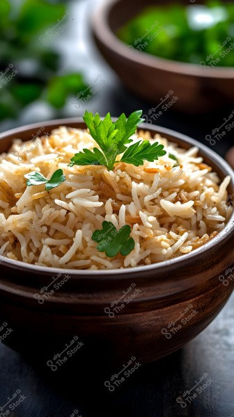Image of a bowl of basmati rice garnished with fresh parsley in a brown wooden bowl. The rice appears perfectly cooked with fried onion bits on top. In the background, another bowl is visible with a blurred effect, enhancing the focus on the main dish.
