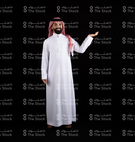 بورتريه رجل سعودي يرتدي الزي السعودي، يرفع ذراعه إلى الأعلى ويفتح كفه يقف على خلفية سوداء، رجل يقوم بعمل حركات بيديه بخلفية سوداء