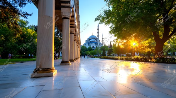 Image depicting an outdoor scene with a large marble column on the left and part of an Ottoman-style mosque in the background, featuring domes and tall minarets. The sun sets behind dense green trees, casting golden rays that filter through the leaves and reflect off the shiny marble floor.