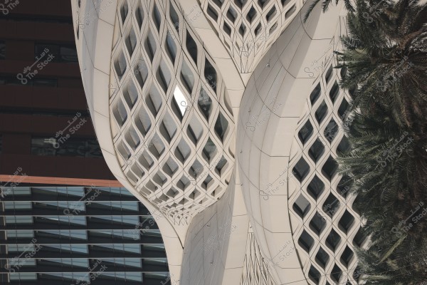 An image of a modern architectural facade featuring a unique, wavy design with interlocking geometric panels that are semi-square. A palm tree is visible on the right, with another building facade in the background.