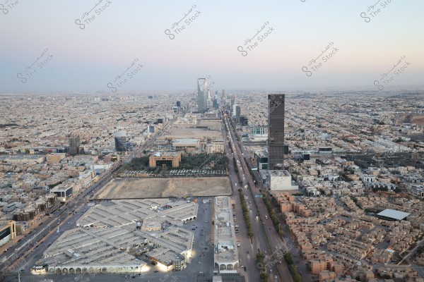 An aerial view of Riyadh city in Saudi Arabia, showing a comprehensive cityscape during the daytime. The Kingdom Tower is visible at the center of the image, surrounded by other buildings, with city streets extending to the horizon.