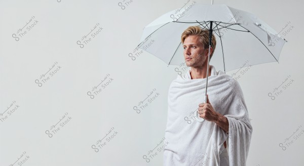 Image of a man wrapped in a white towel holding a white umbrella. He is standing angled to the left, showing only the upper body. The background is white, and he holds the umbrella with his right hand.