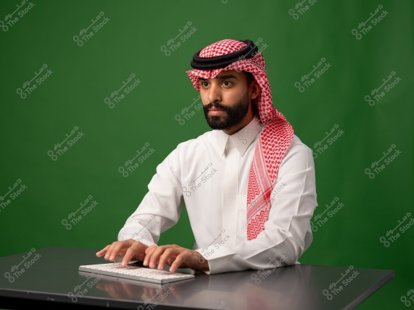 A man wearing a white thobe and a red ghutra sitting at a table using a keyboard.
