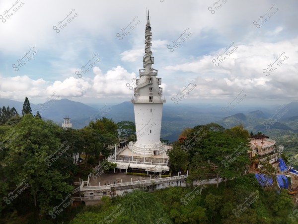 The image shows a tall white tower with spiraling structures, surrounded by lush trees and shrubs. The tower is situated on a green hilltop, and the background features a landscape of green-covered hills with an expansive blue sky dotted with some white clouds.