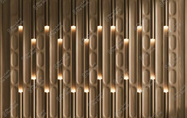 The image shows a wall with a modern geometric design, composed of vertically aligned wooden panels arranged symmetrically. Warm, focused lighting emanates from between the panels, creating an appealing and aesthetic effect on the overall design.