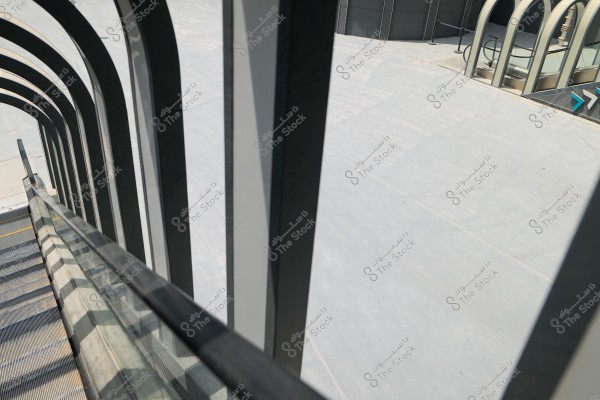 An image showing a scene of an escalator covered in a glass enclosure. The metallic steps are visible on the left with reflections of sunlight on the glass surfaces. In the background, there is a wide cement area with a modern building featuring unique architectural design with curved glass shapes.