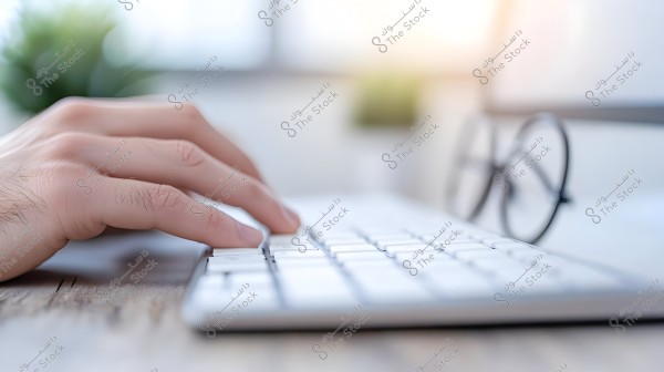 The image shows a hand typing on a white keyboard placed on a wooden table. In the blurred background, there is a pair of glasses, adding a subtle out-of-focus effect. The lighting is soft, giving a calm impression to the scene.