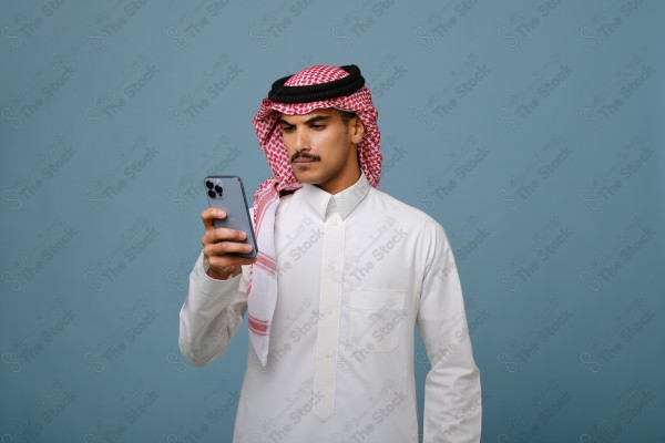 بورتريه شاب سعودي يرتدي الزي السعودي يشير إلى هاتفه ويتفاعل معه، وتظهر على ملامحه الدهشة ، على خلفية سماويه