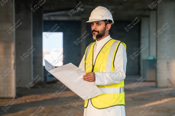 Image of a man wearing a safety helmet and a yellow reflective vest, standing in an unfinished construction site, holding a blueprint. He appears to be wearing traditional clothing that may be from Saudi Arabia.