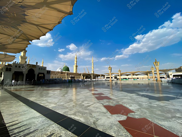 The courtyard of the Prophet\'s Mosque in Medina under a blue sky filled with clouds, with open umbrellas, the minaret, and the green dome in the background.