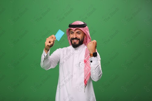 Portrait of an excited Saudi man on a green background, holding a debit card, ready to shop