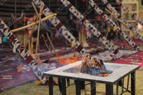 The image depicts a scene inside a traditional Arab tent, featuring a fire burning in a stone fireplace in the foreground. The tent is decorated with colorful rugs and traditional designs in red, black, and white colors, with woven decorations in various colors hanging in the background. The tent reflects traditional Arab culture.