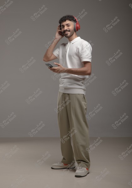 A man wearing red headphones, a white polo shirt, beige pants, and white sneakers stands smiling while holding a mobile phone in hand. He is in a studio with a simple gray background, creating a modern and casual atmosphere.