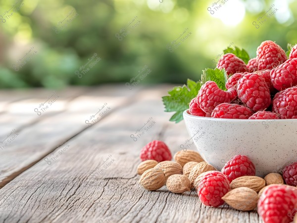 An image of a white bowl filled with fresh red raspberries, placed on an old wooden table outdoors. Surrounding the bowl are several unpeeled almonds. The background features a blurred view of trees and greenery, indicating the photo was taken in a garden or outdoor area.