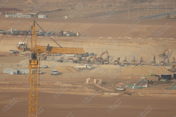 An image of a large construction site in a desert area. The picture shows a large yellow crane and several excavators working on the ground to level the sand and rocks. Trucks and scattered containers are visible around the site. The background features desert terrain with distant roads and several industrial buildings.