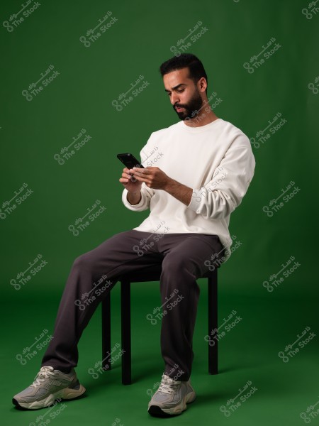 Image of a man sitting on a chair against a green background. The man is wearing a white sweatshirt, gray track pants, and sneakers. He is holding a mobile phone and appears focused on it.