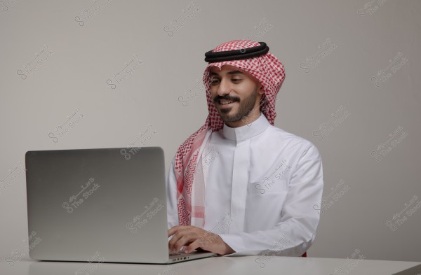 يظهر في الصورة رجل يرتدي الثوب العربي التقليدي والغترة والعقال جالساً أمام جهاز لابتوب. يبدو سعيداً بينما يعمل على الجهاز. الخلفية بيضاء وبسيطة.