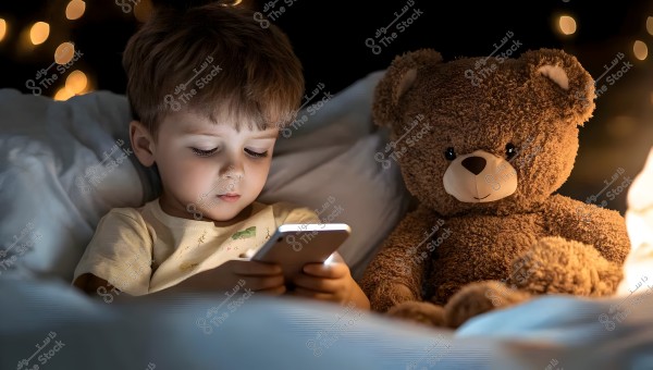A young child sitting in bed, wearing light-colored pajamas, holding a smartphone that emits a bright light. The child is deeply focused on the screen. Beside him is a large brown teddy bear. The background is dark with soft, blurred lights creating a cozy and soothing atmosphere.