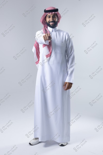 صورة لرجل يرتدي الثوب السعودي التقليدي الأبيض والشماغ الأحمر, يقف مبتسمًا ويوجه إصبعه نحو الكاميرا في استوديو بخلفية بيضاء.