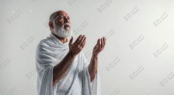An image of an elderly man in a prayer position, wearing a traditional white cloth symbolizing Ihram attire. The background is white, adding a spiritual atmosphere to the image. The man has a reverent expression.