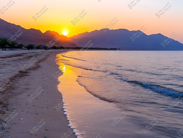 A tranquil beach with the sun setting behind distant mountains. The golden and orange hues of the sun reflect on the water and sand, creating a beautiful scene. A few huts are visible along the beach with sparse green trees on the left, offering a sense of calm and natural beauty.